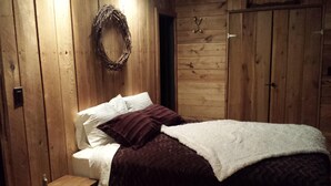 A wooden cabin room with a bed, a small table, and a wall-mounted wreath. - Seven Springs Cabin - Fox Tail Chalet - Authentic, Pet Friendly Log Cabin Chalet (Champion)