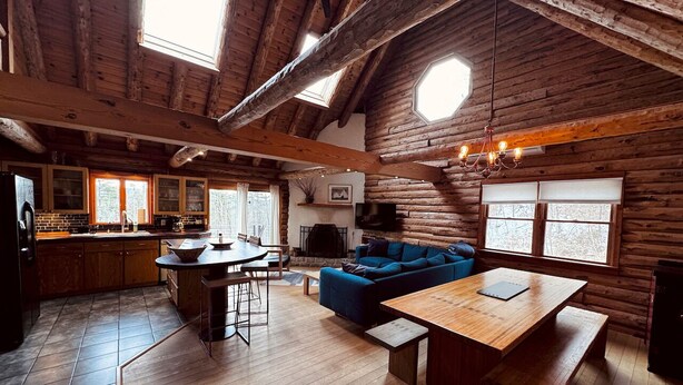 A rustic cabin interior with a wooden dining table, blue sofa, and a kitchen area with wooden cabinets and a large window.