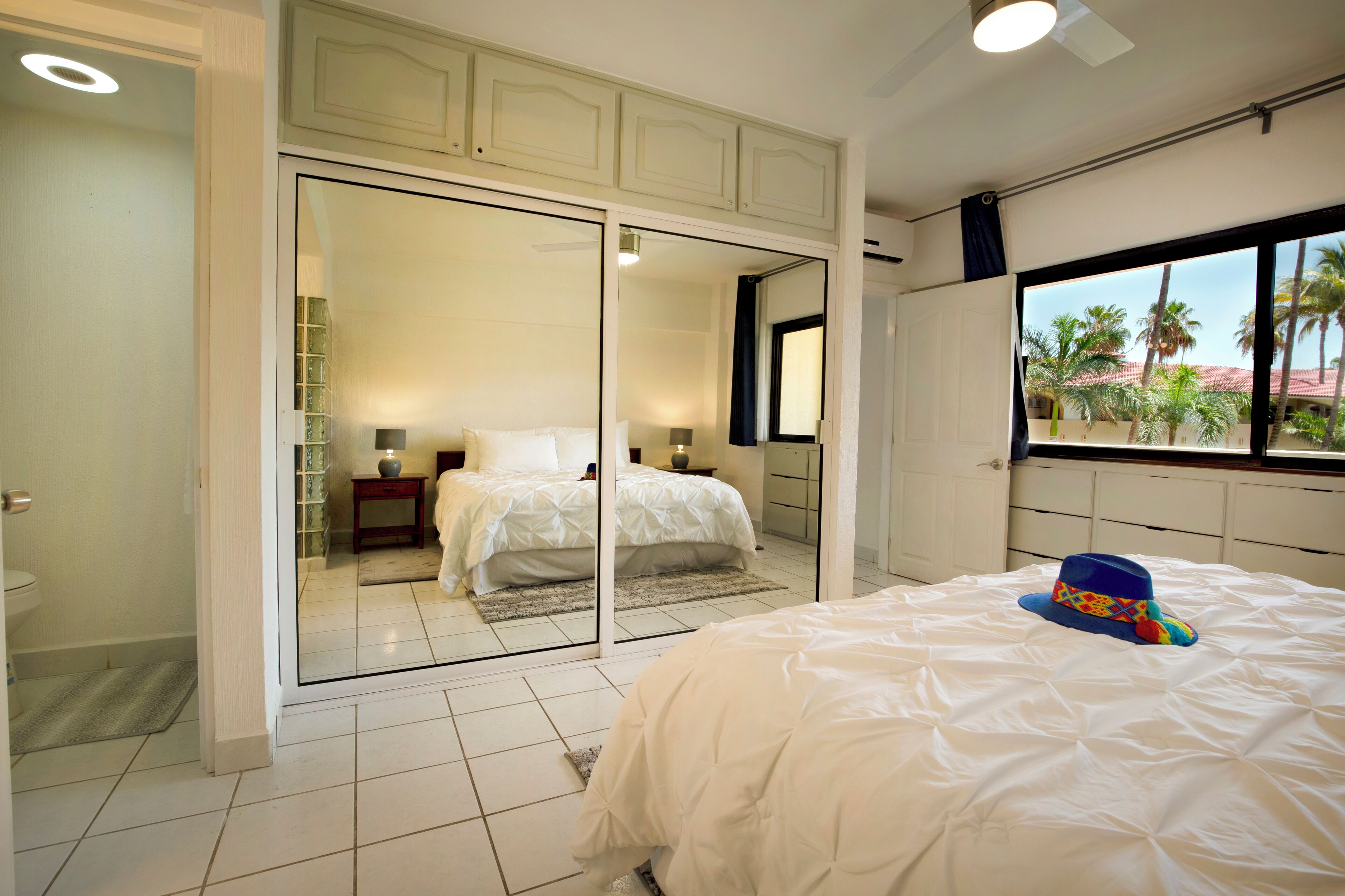 A bedroom with a large bed, a sliding glass door leading to a bathroom, and a view of palm trees outside.