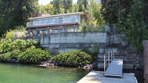 Une propriété au bord du lac, dotée d’un escalier en béton menant à un balcon, d’une jetée en bois s’avançant dans l’eau et d’une végétation luxuriante qui entoure les lieux.