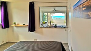 A bedroom with a bed, a window with a view of a balcony, a ceiling fan, and a small shelf with plants. - Grand Case beach (Grand Case)