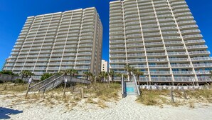 Dos edificios altos con varios balcones, una zona de playa arenosa con una escalera que conduce a los edificios, y un cielo azul despejado.