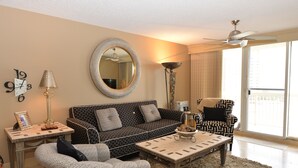 A living room with a patterned sofa, a coffee table, a side table with a lamp, a mirror, and a ceiling fan. - Gulf View, Steps from the Beach, Family Friendly (Destin)