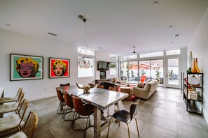 A spacious living area with a dining table, chairs, and a sofa. There are large windows providing natural light and a view of the outdoors. - LUXURY, MODERN DESERT RETREAT 3 BR WITH SALTWATER POOL+SPA  #225624 (La Quinta)