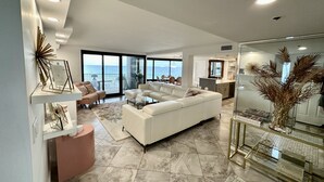 Un salon spacieux avec un grand canapé blanc, une table basse en verre et une vue sur l’océan grâce à de grandes fenêtres.
