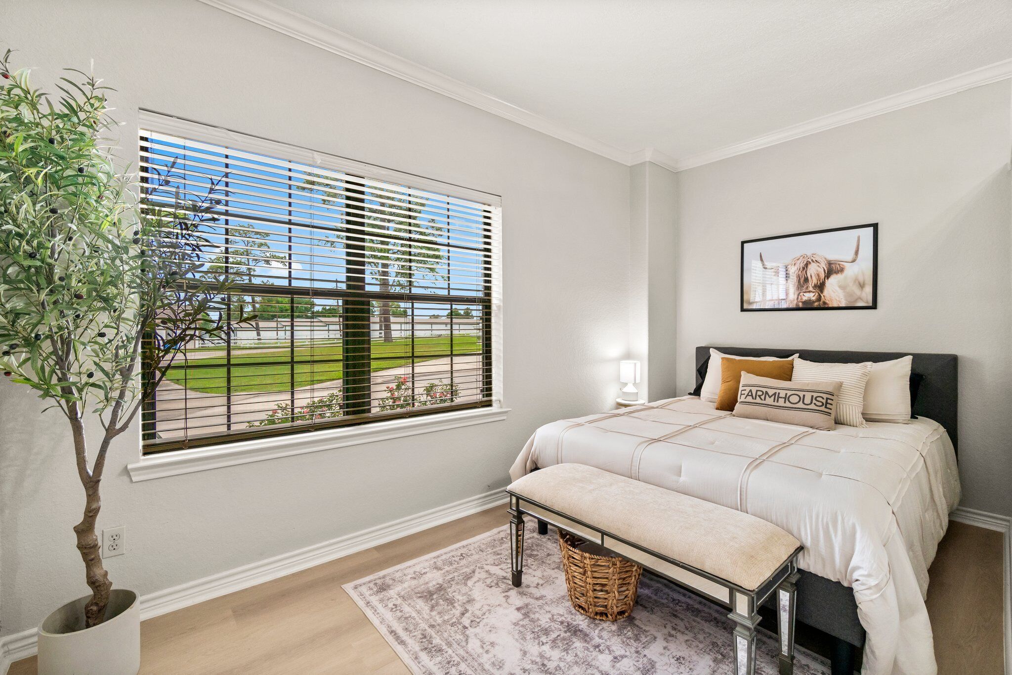 A bedroom with a large window, a bed with a headboard, a bedside table, a lamp, a framed picture on the wall, and a potted plant.