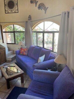 A living room with a purple sofa, a glass coffee table, and a lamp. There are two windows with curtains, and a staircase is visible in the background.
