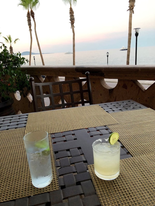 A table with two glasses of beverages, a chair, and a view of the sea with a ship in the distance.
