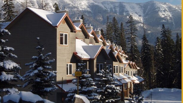 A row of chalet-style buildings with steep roofs, surrounded by snow-covered trees and mountains in the background.