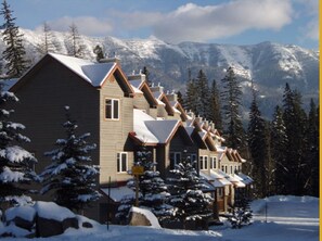 A row of chalet-style buildings with steep roofs, surrounded by snow-covered trees and mountains in the background. - Ski-in Condo Townhouse @Fernie Alpine Resort brand new Hot Tub summer 2026 & BBQ (Fernie)