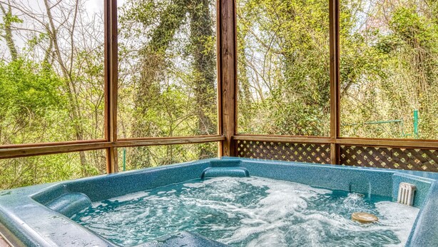A hot tub with a wooden deck and glass walls, surrounded by lush greenery. - River View Rental-Gameroom-Only 1 Mile Off Main Parkway Pigeon Forge-Hot Tub and more (PIGEON FORGE)