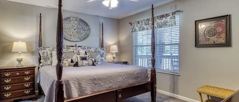 A four-poster bed with a ceiling fan, a large decorative plate, and a window with blinds.