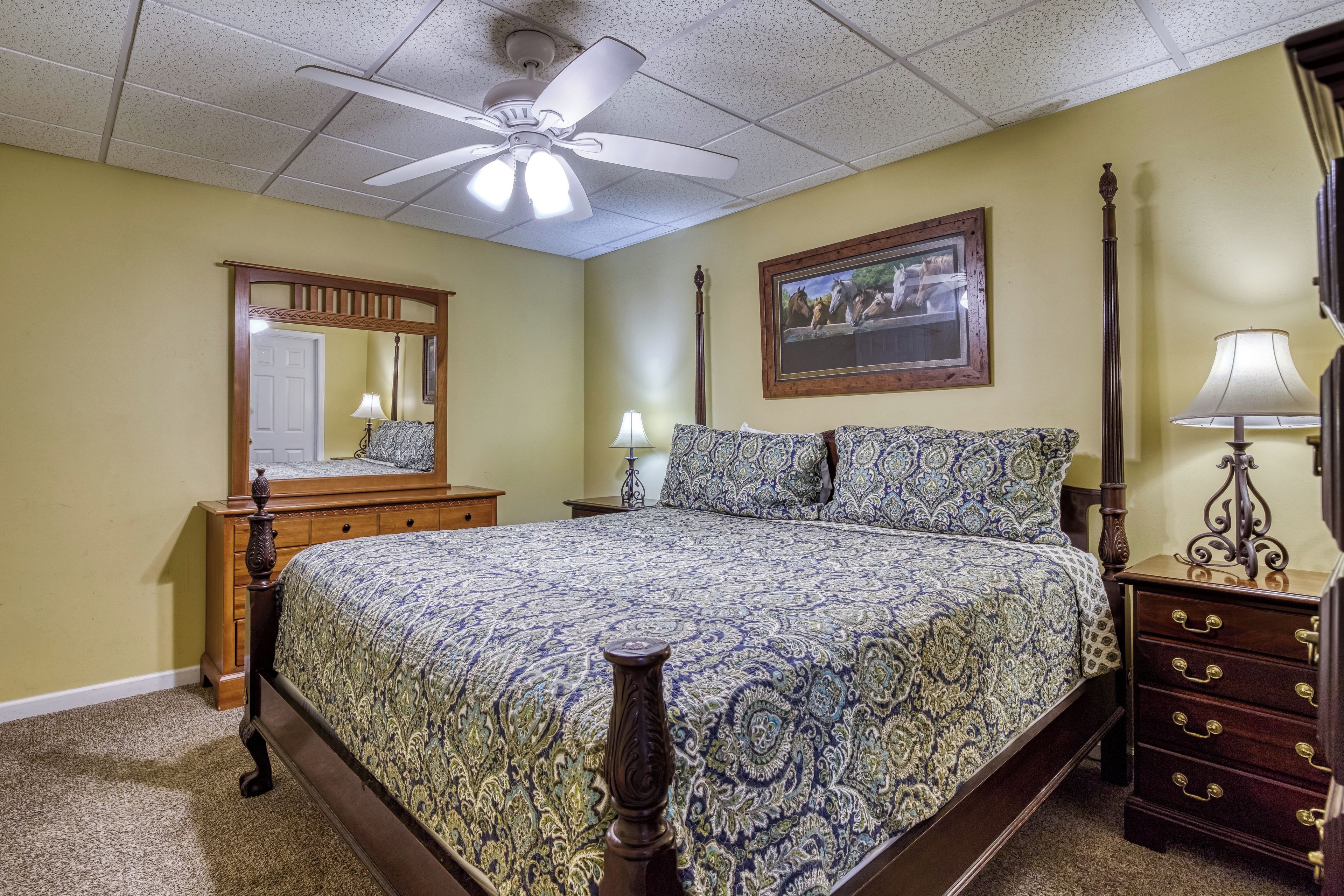 A bedroom with a bed, a dresser, a nightstand, a lamp, a ceiling fan, a mirror, and a framed picture on the wall.