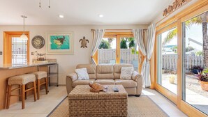 A living room with a sofa, ottoman, and bar stools. There is a painting on the wall, a clock, and a window with curtains. - Lazy Tortuga ground level 2bed/2bath half duplex with dockage & cabana club (Key Colony)