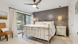 Une chambre à coucher avec un lit en métal, une table de chevet blanche, une chaise en osier et un ventilateur de plafond.