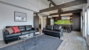 Ein modernes Wohnzimmer mit einem schwarzen Ledersofa, einem Glastisch und einer Küche im Hintergrund, deren Rückwand grün ist.