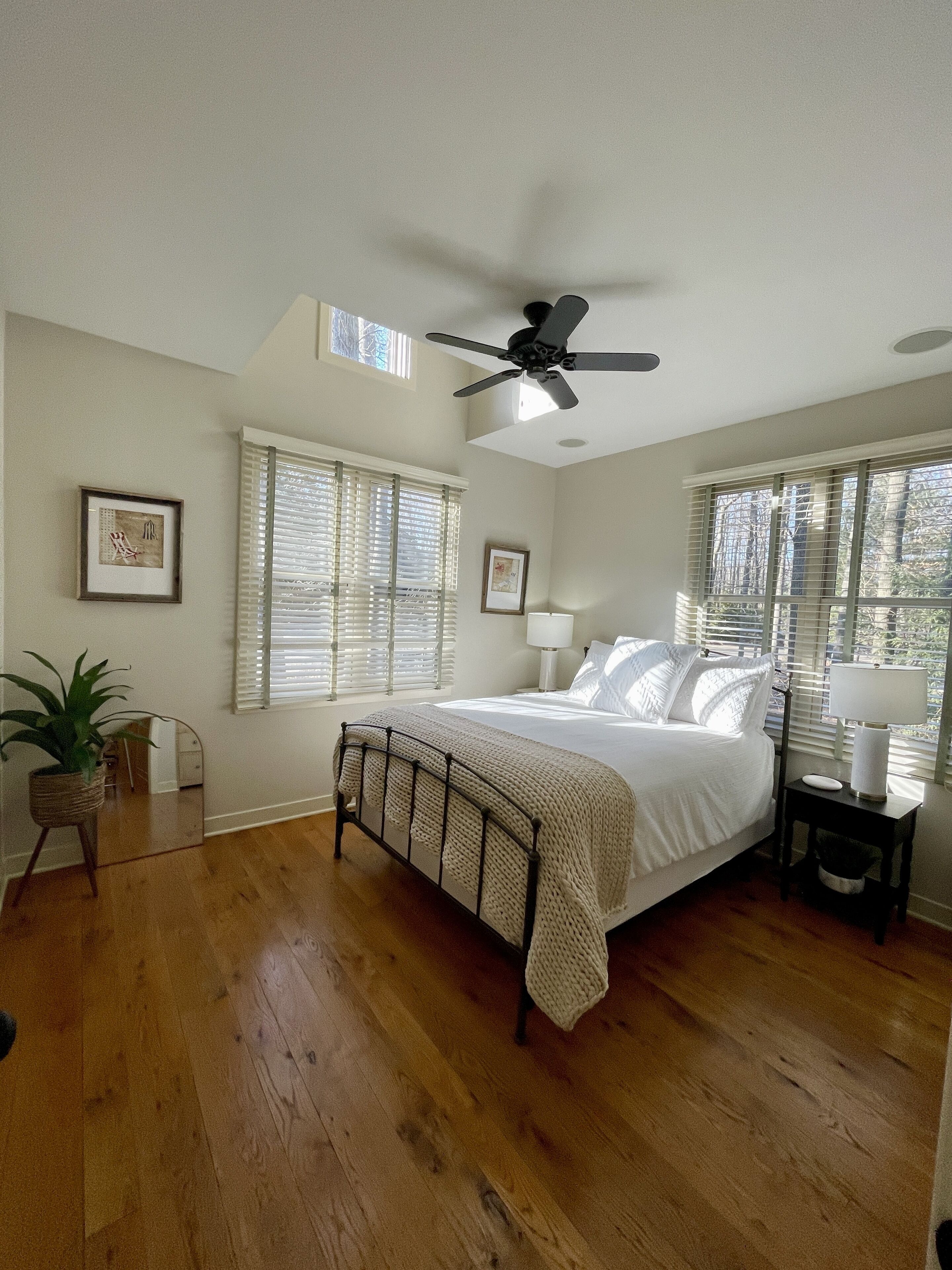 Une chambre à coucher avec un lit, un ventilateur de plafond, une plante en pot et une fenêtre avec des stores.