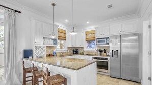 A modern kitchen with white cabinetry, a central island with bar stools, stainless steel appliances, and a large window with blinds. - Sunset Seeker-Gulf Side 30A Villa w/ Community Pool,Pet Friendly! (Seacrest Beach)