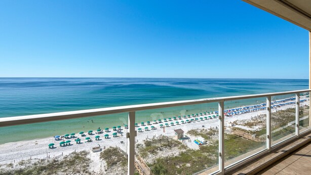 Une vue sur la plage depuis un balcon, avec un ciel bleu dégagé, une mer turquoise et des rangées de parasols de plage.