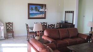 A living room with a brown leather sofa, wooden dining table, chairs, and a painting of a beach scene. - PRIVATE LUXURY Beachfront Home, Beach Cabana, Kayaks, Paddle Board (Casuarina Point)
