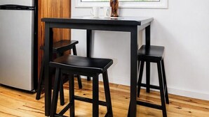 A kitchen with wooden cabinets, a stainless steel refrigerator, a black table, two black stools, and a window with a view of greenery. - #S4-West Bay 1 Bed Apt - 2 min walk to the beach! (West Bay)