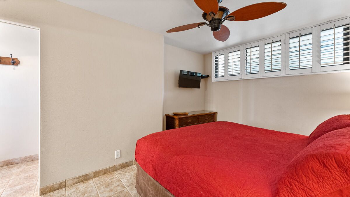 A bedroom with a bed, a ceiling fan, a TV mounted on the wall, and a window with blinds.