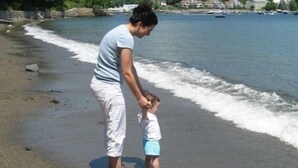 Una mujer y un niño en una playa arenosa con olas, cielo despejado y una montaña al fondo.