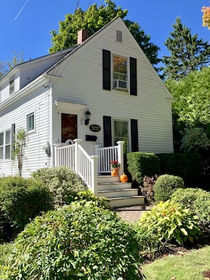 A white two-story house with black shutters, a white porch, and a small garden with plants and pumpkins.