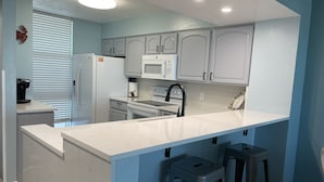 A modern kitchen with white cabinets, a white countertop, a built-in microwave, and a coffee maker. There are two stools and a window with blinds. - Panama City Beachfront! 5STAR Dog Friendly! Vacation-Staycation or MicroCation! (Panama City Beach)