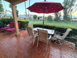 Une terrasse avec une table et des chaises, un parasol rouge, et une vue sur une pelouse verdoyante et des bâtiments.