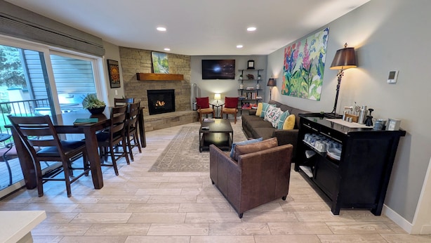 A spacious living room with a fireplace, a dining area with wooden chairs, a sofa, and a coffee table. There is a TV mounted on the wall and a painting above it. - Luxury Condo w/ FREE WiFi, Parking, Heated Pool, Hot Tubs, Skier Shuttles (Avon)