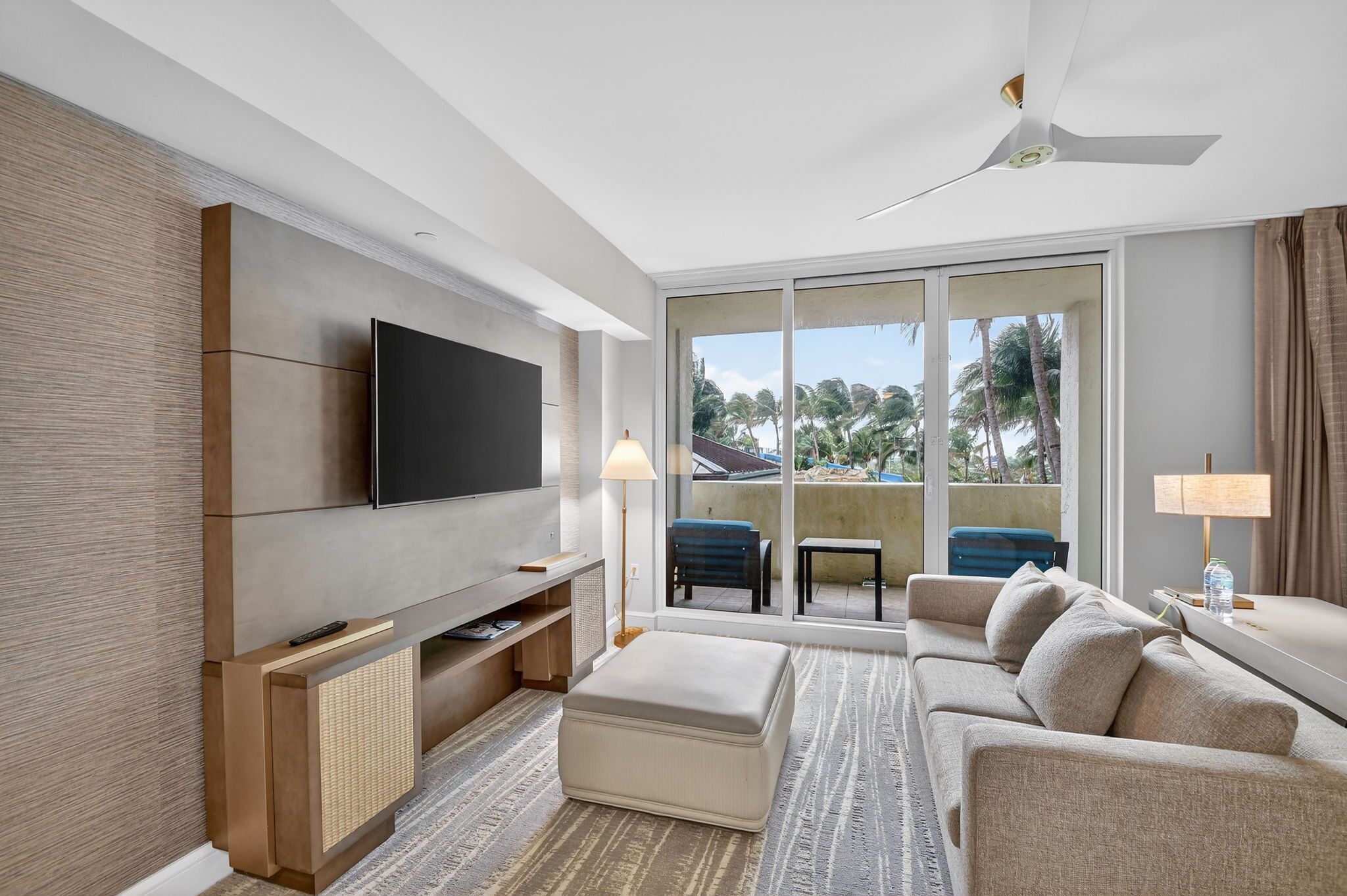 Een moderne hotelkamer met een flatscreen-televisie, een bank, een poef, een kleine tafel en een balkon met uitzicht op palmbomen.