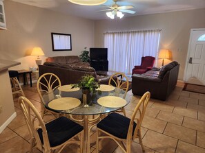 Een woonkamer met een eettafel met glazen blad, omringd door rieten stoelen, een bruine leren bank, een rode fauteuil en een televisie.
