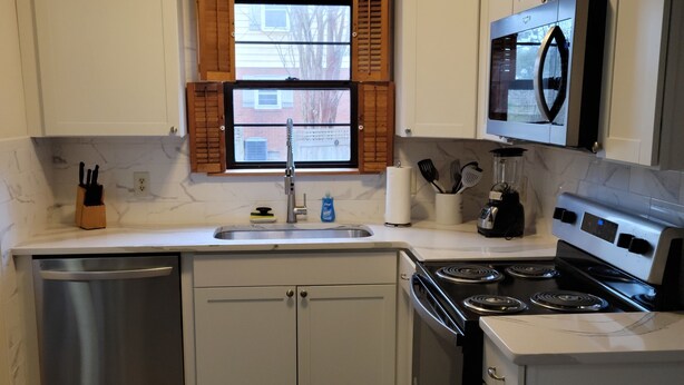 Cocina compacta con armarios blancos, lavavajillas de acero inoxidable, estufa y microondas.