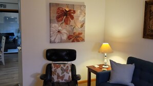 Un salón con un sofá azul, un sillón de cuero negro, una mesa de centro de madera, una lámpara y un cuadro floral en la pared.