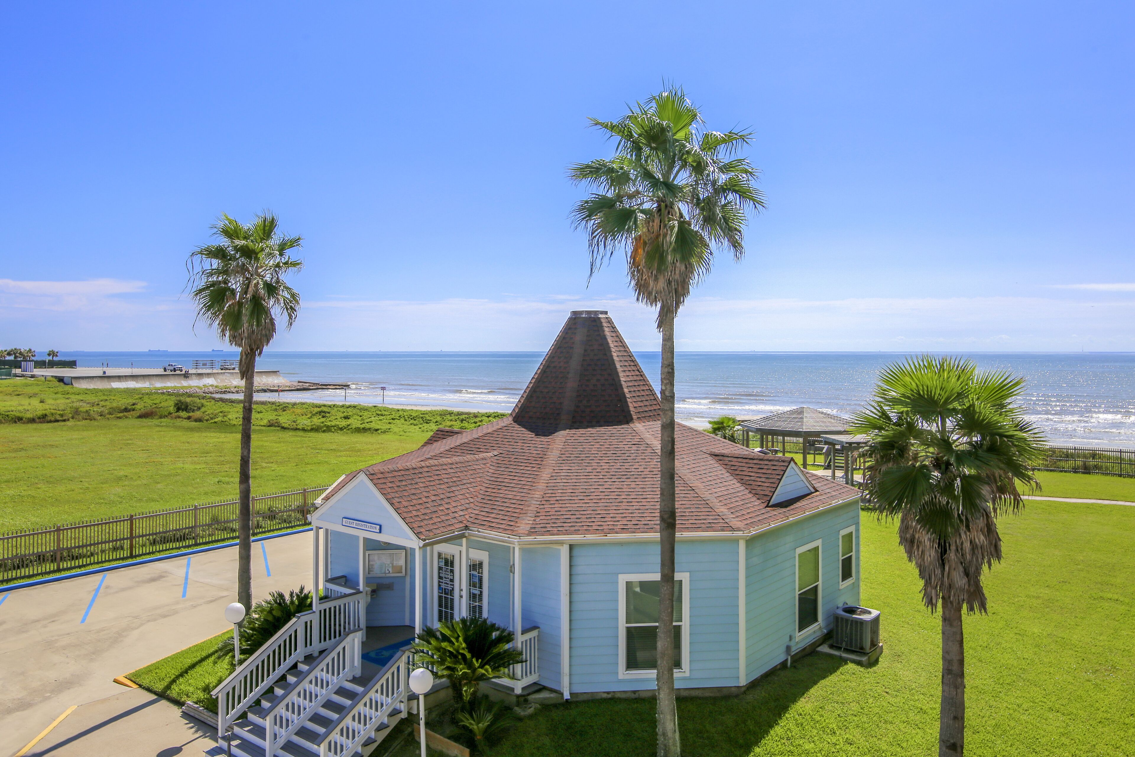 A small, light blue building with a steep roof, surrounded by a well-maintained lawn and palm trees, overlooking a beach.