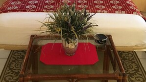 A bedroom with a bed, a painting, a red patterned bedspread, a wooden nightstand, and a glass vase with flowers.