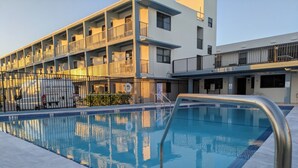 Una piscina con superficie de agua azul cristalina, rodeada por una solana con baldosas. Al fondo, se observa un edificio de varios pisos con balcones y una furgoneta blanca estacionada al frente.