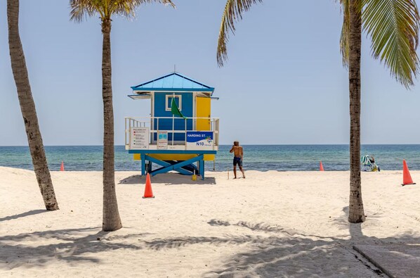 A lifeguard tower with a blue roof and yellow and blue walls, located on a sandy beach with palm trees and the ocean in the background.