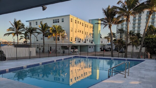 Piscina rectangular con una escalera de metal, rodeada por una cerca negra y palmeras.