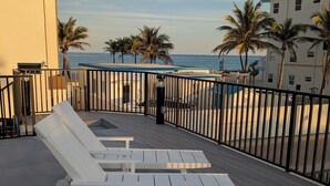 A rooftop terrace with white lounge chairs, a view of the ocean, palm trees, and a building with a balcony.