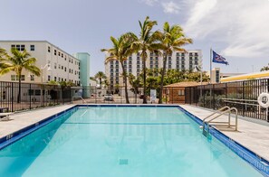 A swimming pool with a clear blue water, surrounded by a black fence, palm trees, and a hotel building in the background. - My Hollywood Beach Rentals B105.. Steps to Beach. Pool and Free Parking (Hollywood)