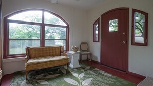 Een kamer met een groot rondboogvenster, een houten bankje, een klein tafeltje en een rode deur.