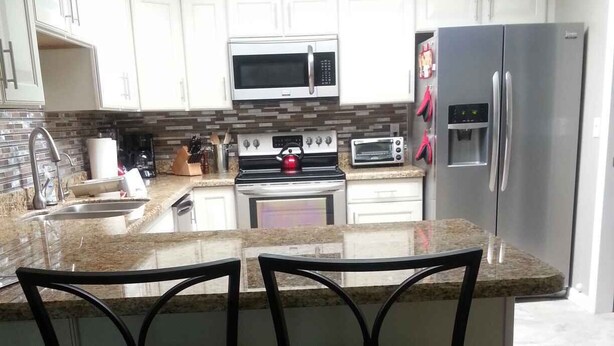A kitchen with white cabinets, a stainless steel refrigerator, a microwave, an oven, and a toaster. There are two black bar stools at the island. - North Central Phoenix - Two Master Stes. 1500 Sf Newly Remodeled, Peaceful!! (Phoenix)