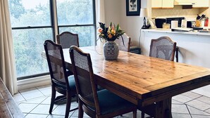 Un coin repas avec une table et des chaises en bois, un vase rempli de fleurs et une vue sur les arbres à travers de grandes fenêtres.