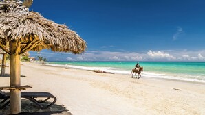 Una playa con un paraguas de paja, sillas reclinables y una persona montando a caballo.