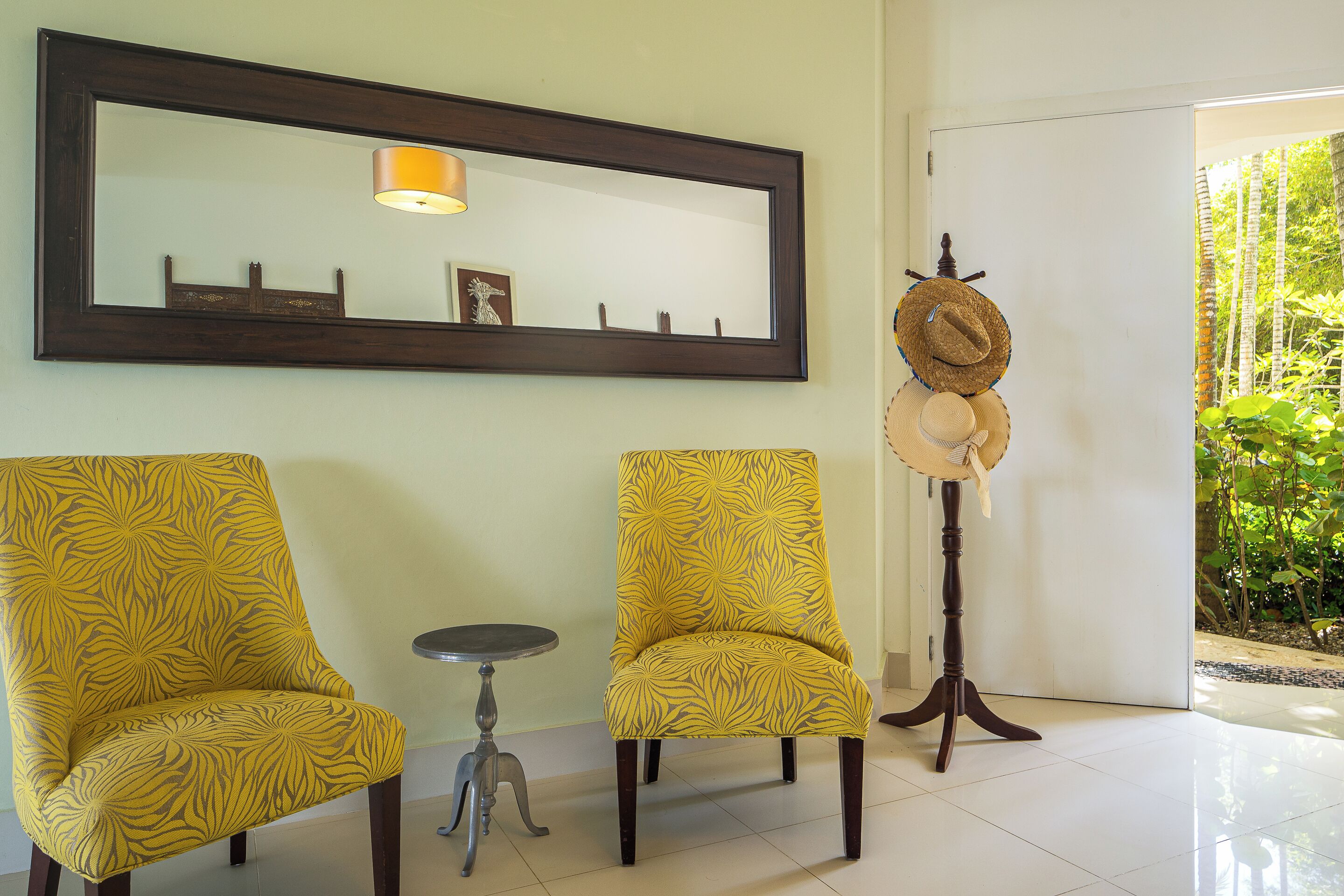 Una habitación con dos sillas amarillas estampadas, una mesita redonda pequeña, un espejo de marco oscuro y un perchero con un sombrero de paja.