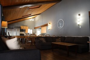 A modern living room with a dark sofa, wooden coffee table, and a dining area with wooden chairs and a kitchen in the background. - Spacious 11BR Cabin for Groups | Near Granby Ranch (Granby)