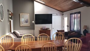 Un coin repas avec une table et des chaises en bois, un téléviseur fixé au mur et un canapé à l’arrière-plan.