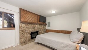 A bedroom with a stone fireplace, a bed with a quilted cover, a bedside table with a lamp, and a window with curtains.